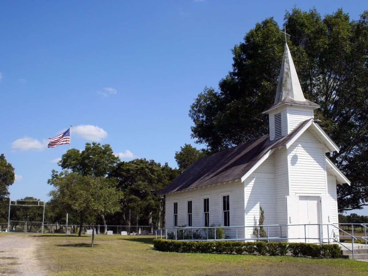 A small rural church in Texas. There is a cemetary and a large oak tree behind the church. - Texas View