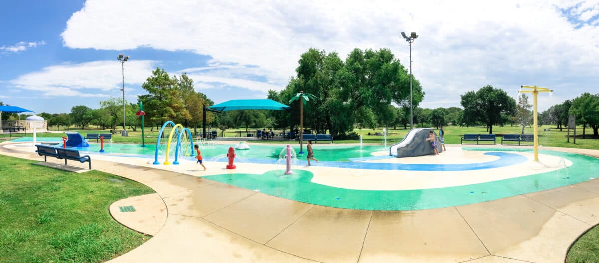 Panorama view rear view Asian toddler boy playing at splash park near Dallas Texas America. Colorful recreation site with splashing water fountains for kids summertime activities. - Texas View