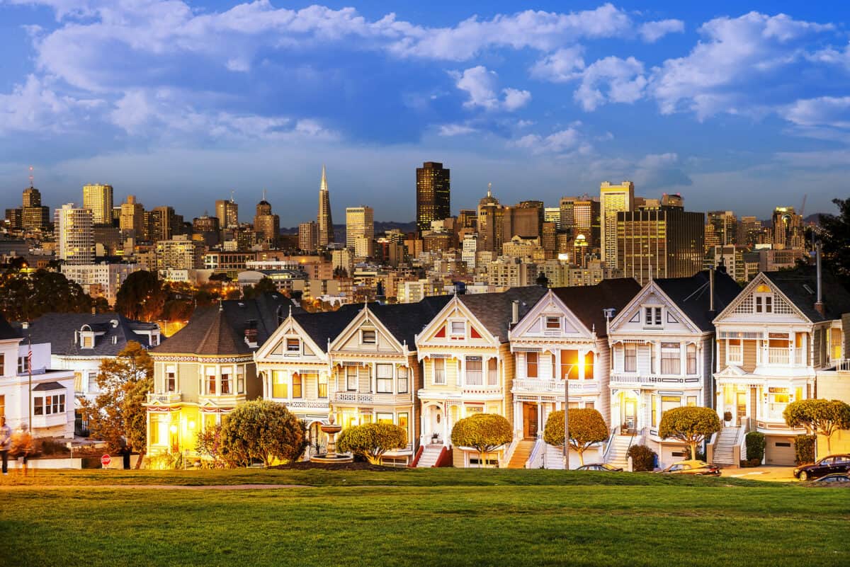 The Painted Ladies of San Francisco California sit glowing amid the backdrop of a sunset and skyscrapers. - Texas View