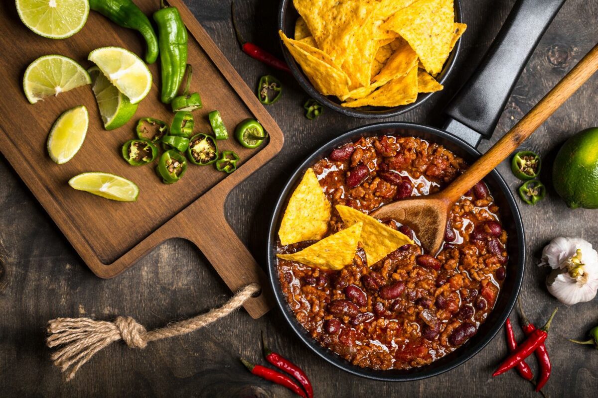 Chili con carne in frying pan on dark wooden background. Ingredients for making Chili con carne. Top view. Chili with meat nachos lime hot pepper. MexicanTexas traditional dish Chili con carne - Texas News, Places, Food, Recreation, and Life.
