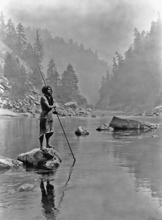 A smoky day at the Sugar Bowl--Hupa. Hupa man with spear, standing on rock midstream, in background, fog partially obscures trees on mountainsides. Published 1924 in The North American Indian / Edward S. Curtis. [Seattle, Wash.] : Edward S. Curtis, 1907-30, Suppl., v. 13, pl. 471.
