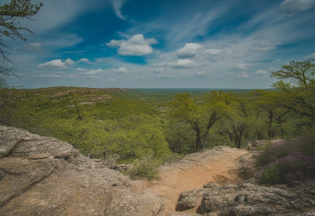 A scenic hiking trail winding through rocky hills and green trees under a blue sky with clouds.