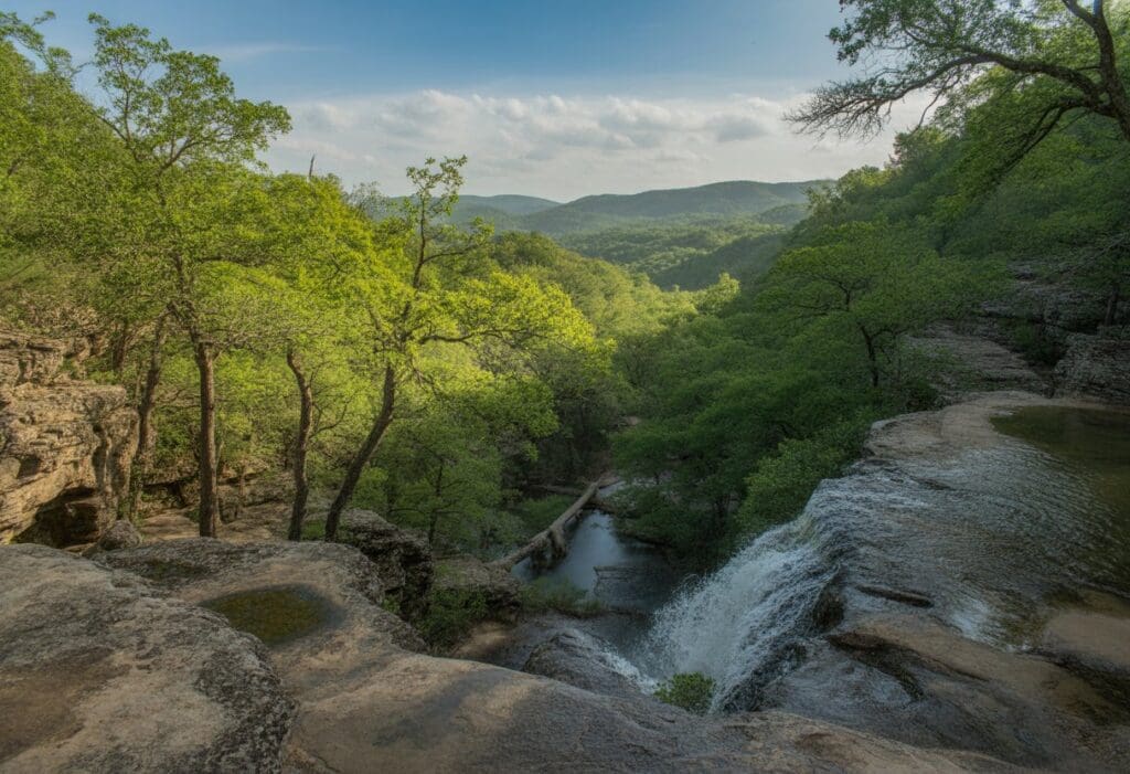 A scenic view of waterfalls, mountains, and forest with a hiking trail winding through the trees.