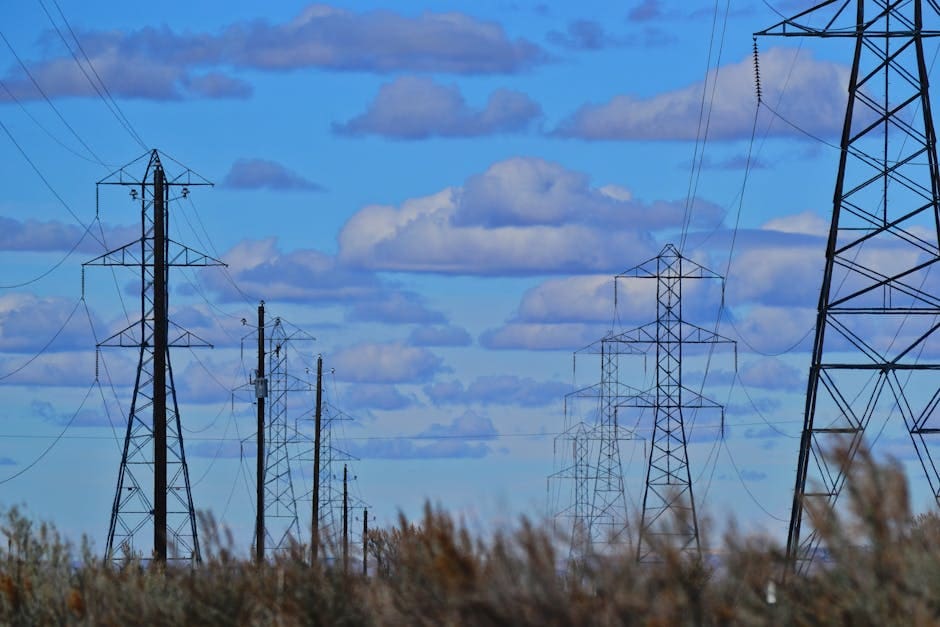 Transmission towers and power lines silhouetted against a bright blue sky with clouds.