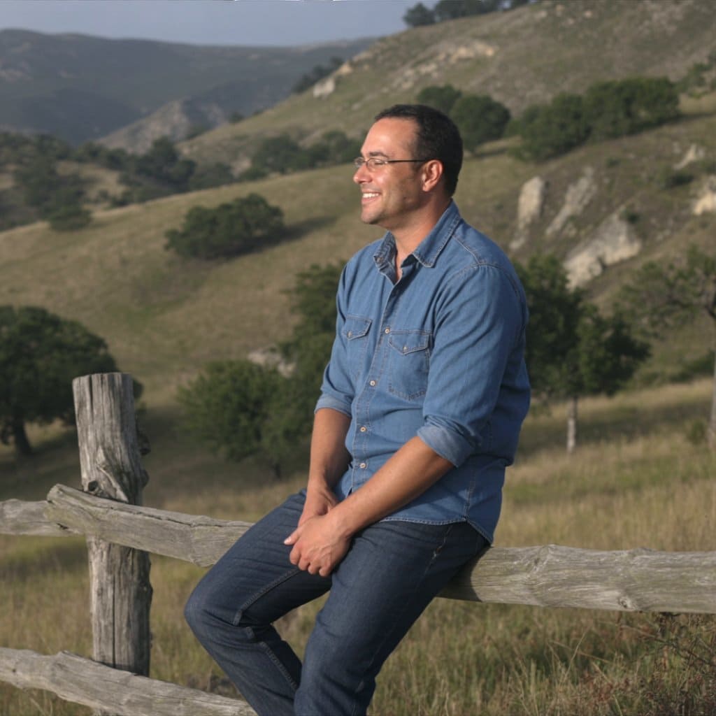 Christian Linden sitting in a Texas field, smiling. Texasview.com shows Linden in a denim shirt, enjoying the Texas landscape and sunshine.